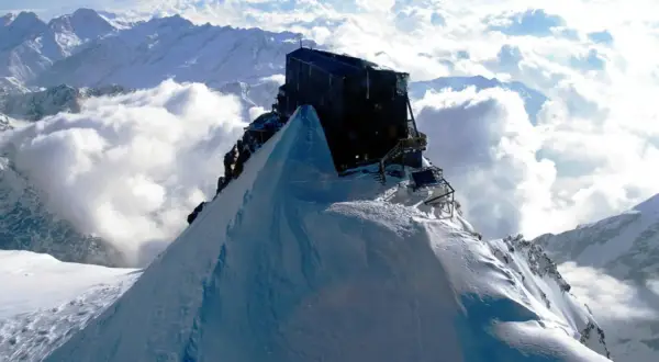 Il rifugio più alto d'Europa, un edificio scuro su una vetta innevata, domina un mare di nuvole e montagne.