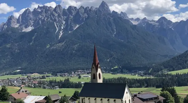 Chiesa con campanile rosso in primo piano, sullo sfondo le maestose vette delle Dolomiti e i **paesi più belli** della **Val Pusteria**.