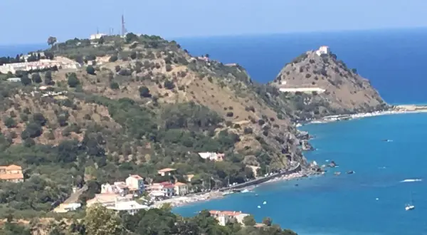 Panoramica di Barcellona Pozzo di Gotto con case bianche e rosse affacciate sul mare blu intenso, colline verdi e un castello in cima.