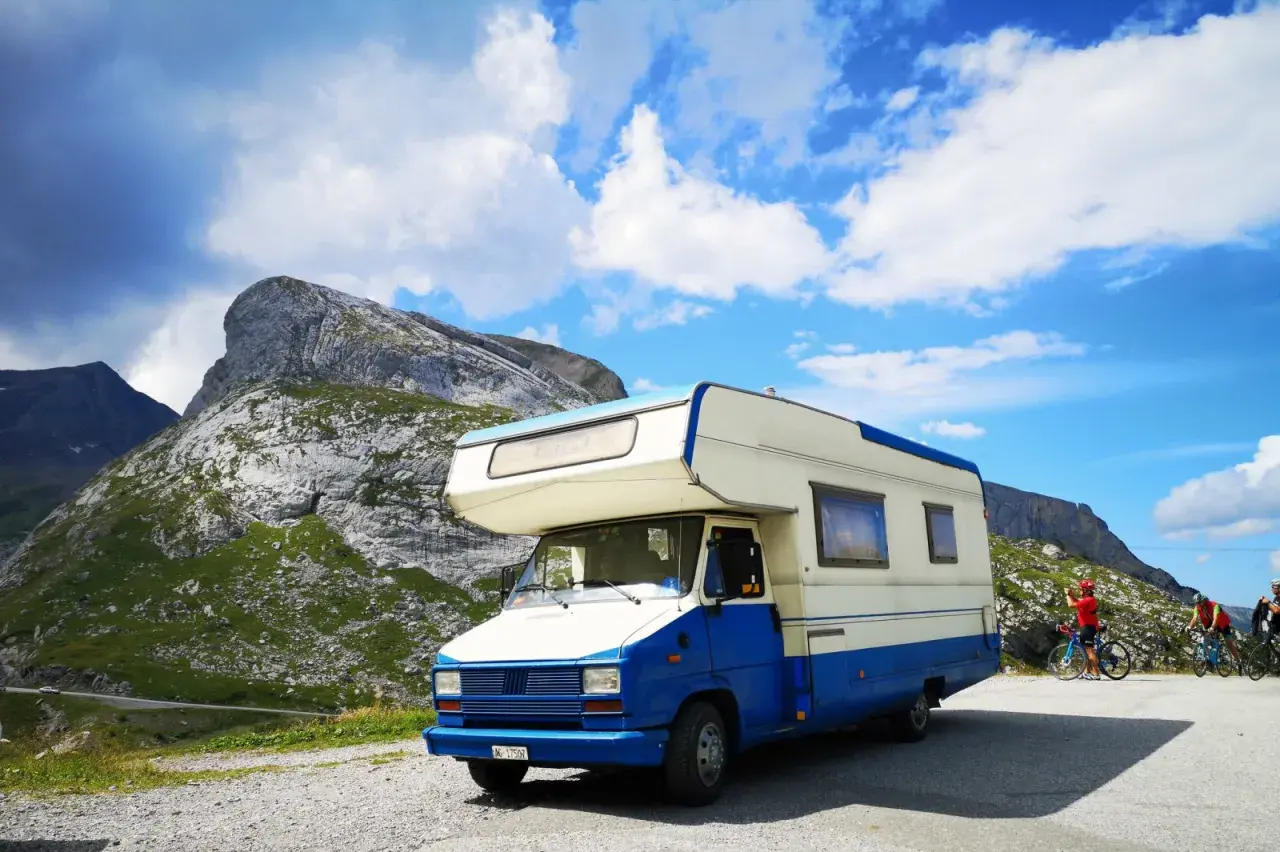 Camper blu e bianco parcheggiato in montagna, pronto per esplorare gli **itinerari camper Italia**. Due ciclisti sullo sfondo.
