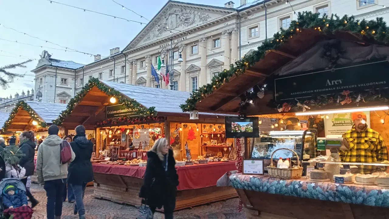 Mercatini di Natale Aosta: bancarelle addobbate con luci e ghirlande, prodotti tipici e gente che passeggia. Atmosfera magica.