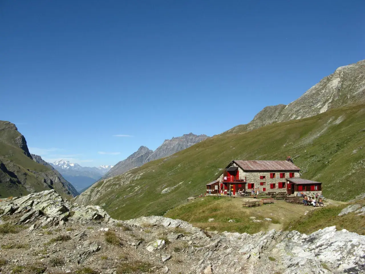 Rifugio nel Val di Rhemes, con persone sedute ai tavoli all'esterno, circondato da verdi pendii montuosi sotto un cielo azzurro.