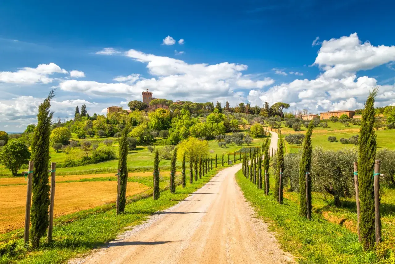 Strada sterrata fiancheggiata da cipressi in una gita in Toscana, con un borgo in collina sotto un cielo azzurro e nuvoloso.