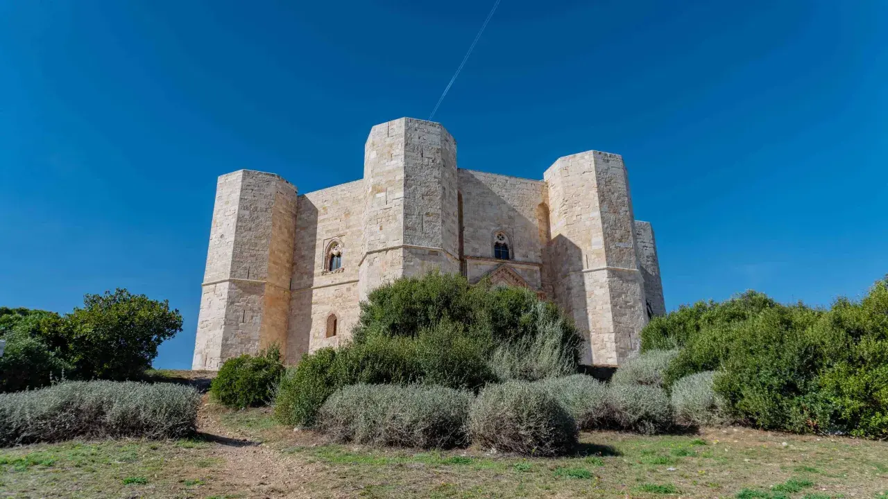 Castel del Monte, un'imponente fortezza ottagonale in pietra chiara, si erge maestoso sotto un cielo azzurro intenso, circondato da vegetazione mediterranea.