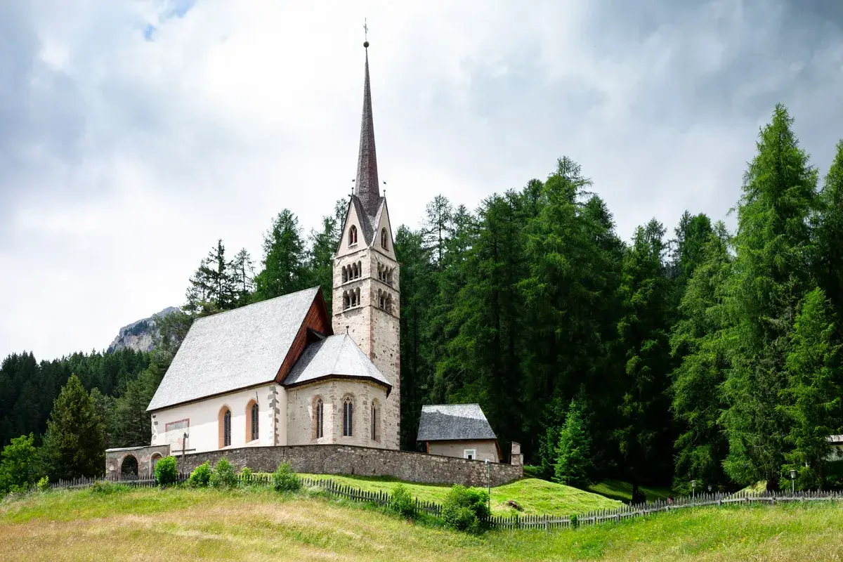 Chiesa di San Giovanni di Fassa, con campanile a cuspide, circondata da un fitto bosco di abeti e montagne sullo sfondo.