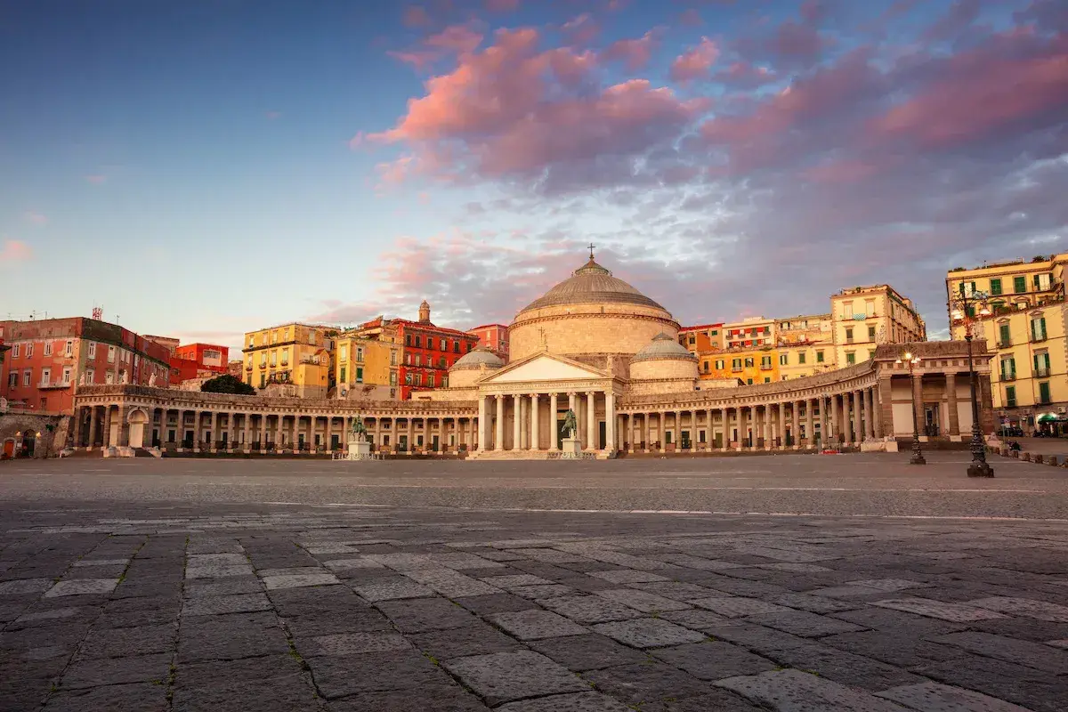Piazza del Plebiscito a Napoli, con il Palazzo Reale e la Basilica di San Francesco di Paola, sotto un cielo rosato al tramonto.