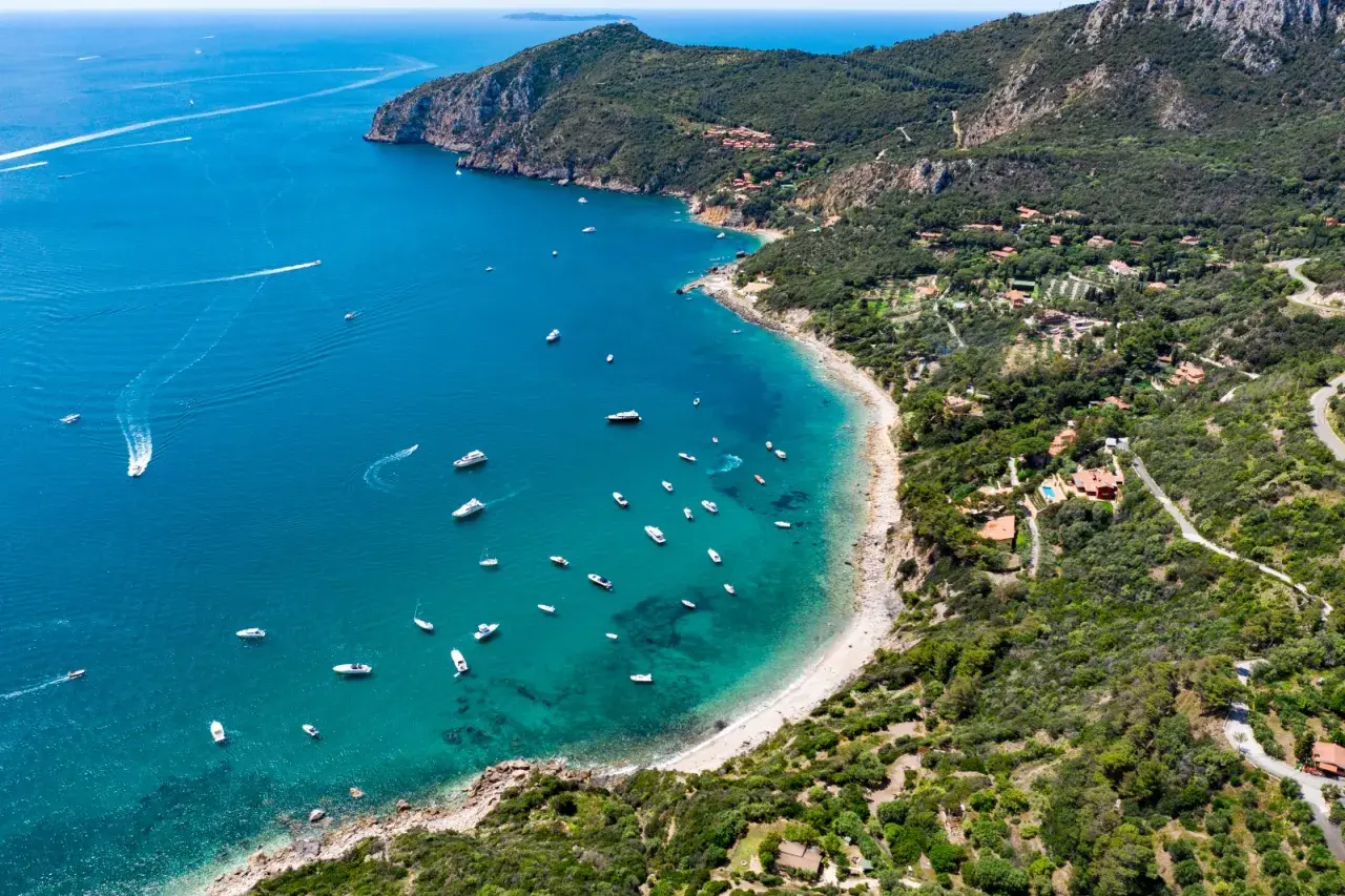 Vista aerea di una baia con spiagge dorate e acque cristalline, un vero gioiello della cartina della Toscana. Diverse imbarcazioni solcano il mare blu intenso.