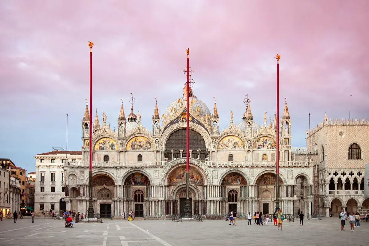 La maestosa Basilica di San Marco a Venezia, con le sue cupole dorate e mosaici scintillanti, si staglia contro un cielo rosa al tramonto.