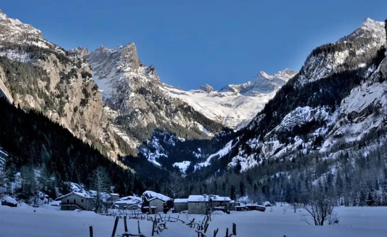 Case inneggiate nel Val di Mello inverno, con montagne imponenti e case di pietra sotto un cielo azzurro.