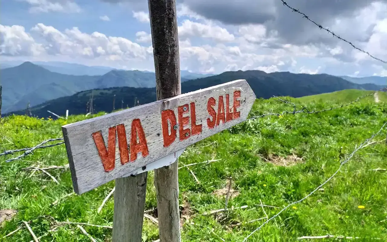 Cartello in legno indica il **via del sale cammino**. Panorami montani verdi e cielo azzurro con nuvole bianche.