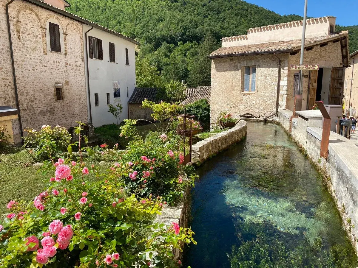 Un ruscello attraversa un borgo umbro, tra case in pietra e rose rosa. Un angolo incantevole tra i borghi più belli dell'Umbria.