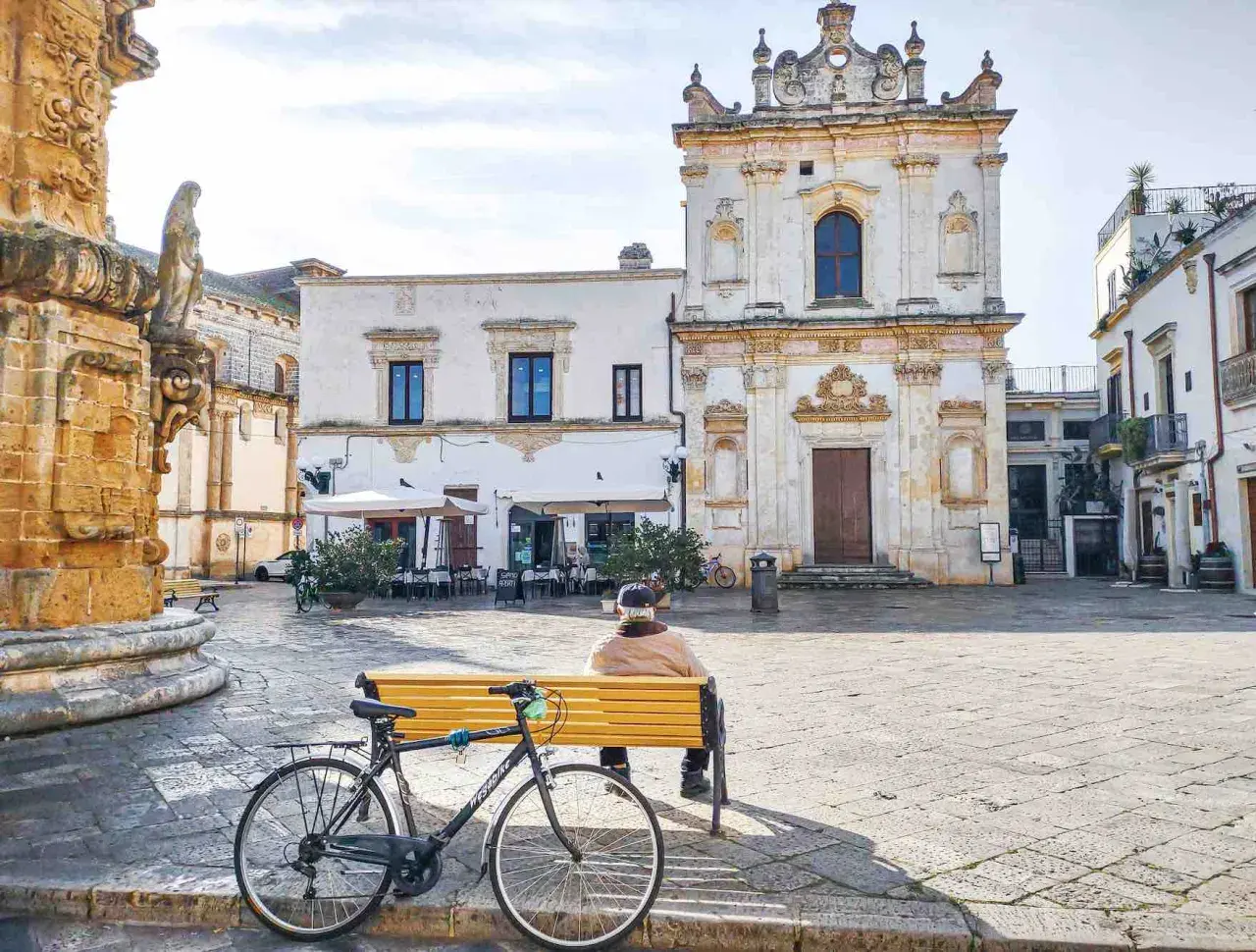 Un uomo seduto su una panchina in una piazza, con una bicicletta accanto. Architettura barocca e scorci dei borghi più belli del Salento.