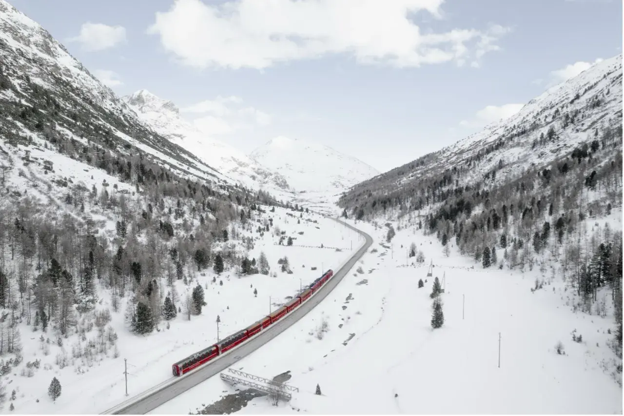 Un treno rosso attraversa una valle innevata, un viaggio tra i **treni panoramici Italia** con montagne maestose e alberi spogli.