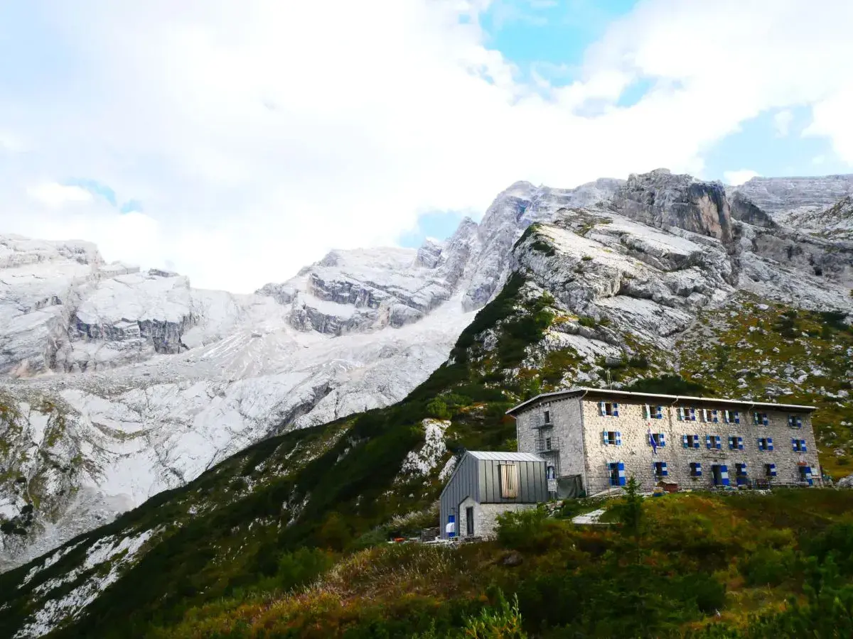 Un rifugio in pietra con persiane blu si staglia su un pendio verdeggiante, sotto imponenti montagne rocciose. Un esempio di rifugi in Veneto.
