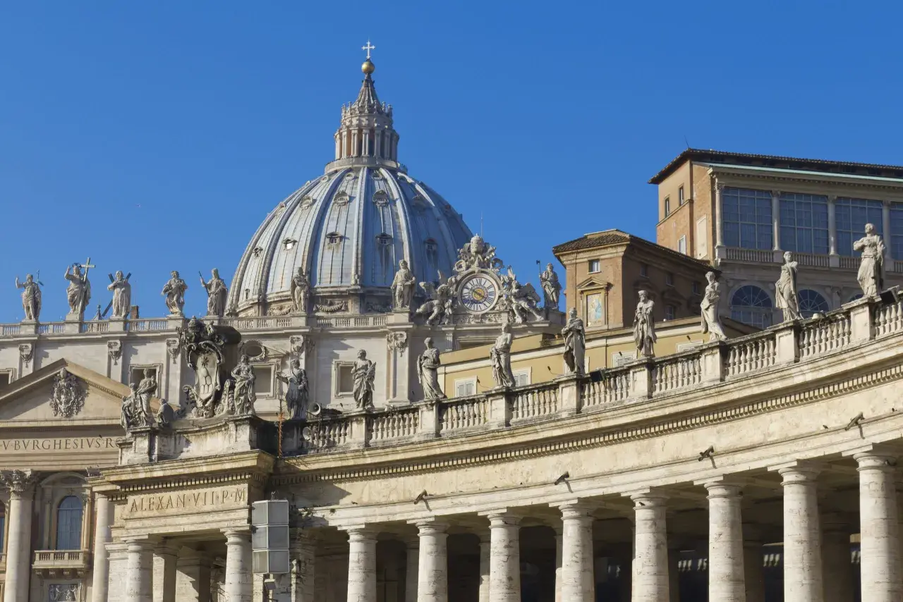 Cupola maestosa e colonnato di San Pietro, una delle quattro basiliche di Roma, con statue che vegliano sul cielo azzurro.