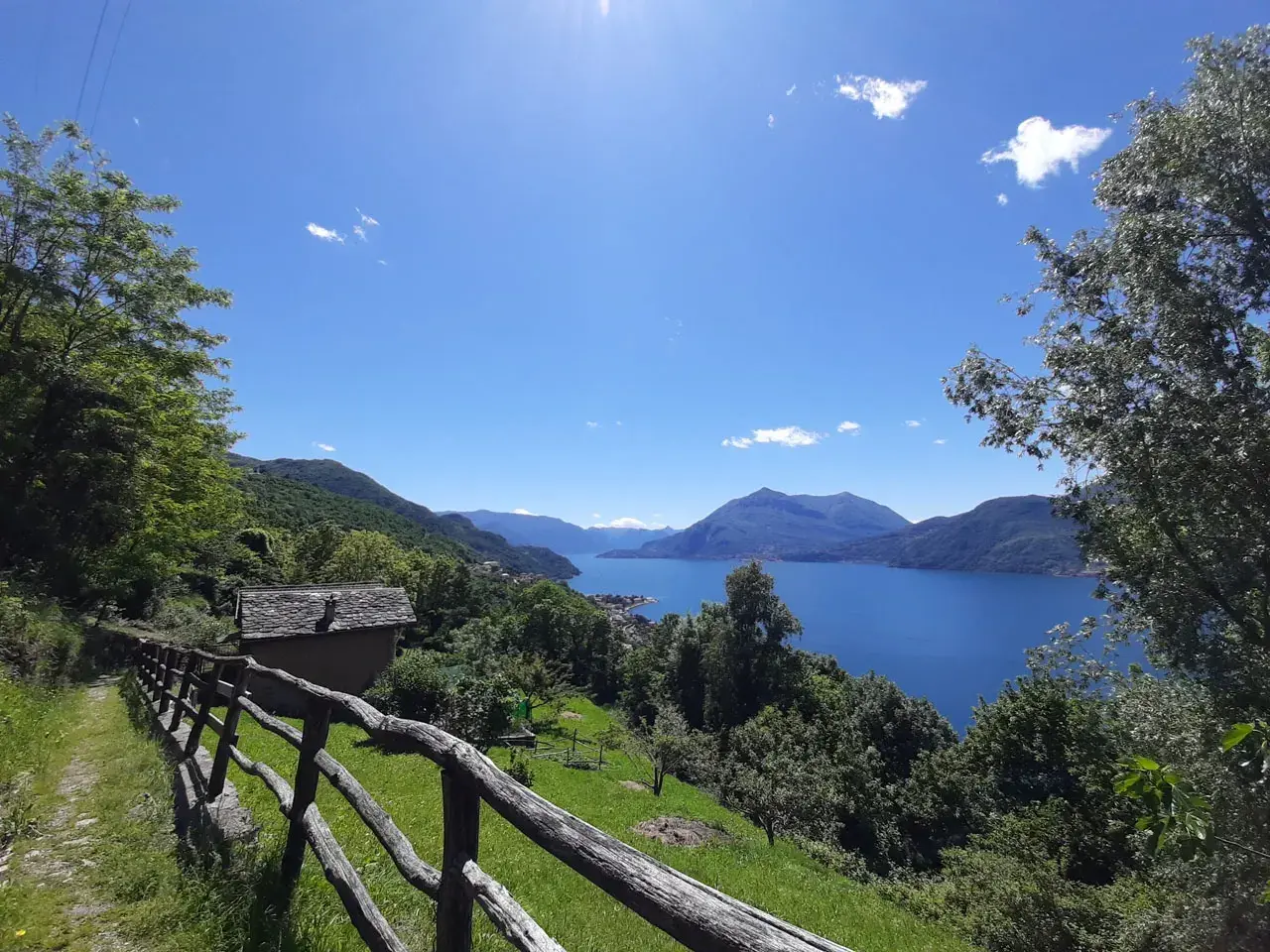Il sentiero del viandante si snoda tra alberi verdi, con una vista mozzafiato su un lago azzurro e montagne sullo sfondo. Una piccola casa con tetto in pietra si intravede.