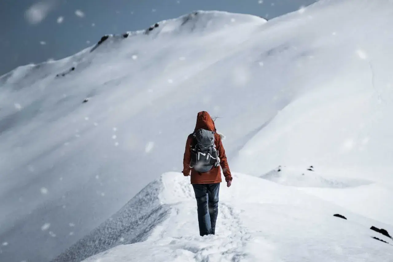 Persona con zaino cammina su un crinale innevato, mentre fiocchi di neve cadono dolcemente.
