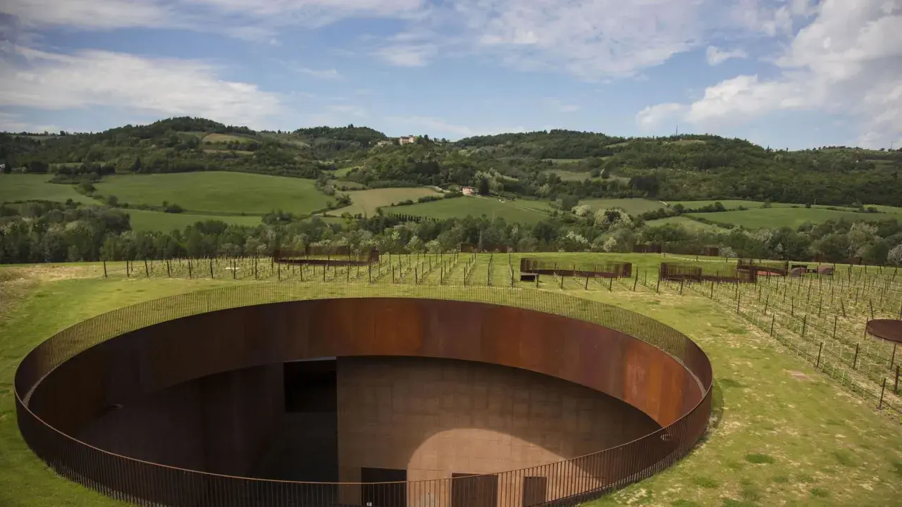 Un cerchio di metallo arrugginito emerge dal verde delle colline del Chianti, con vigneti che si estendono all'orizzonte sotto un cielo azzurro.