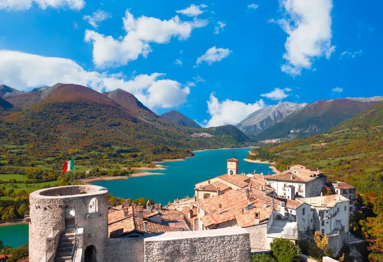Borgo medievale con torre e lago turchese, un incantevole scorcio di cosa vedere in Abruzzo, tra montagne autunnali e cielo azzurro.
