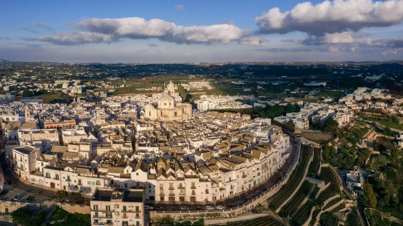 Vista aerea di Locorotondo di sera, con le sue case bianche a tetto spiovente e la chiesa principale che domina il paesaggio collinare.