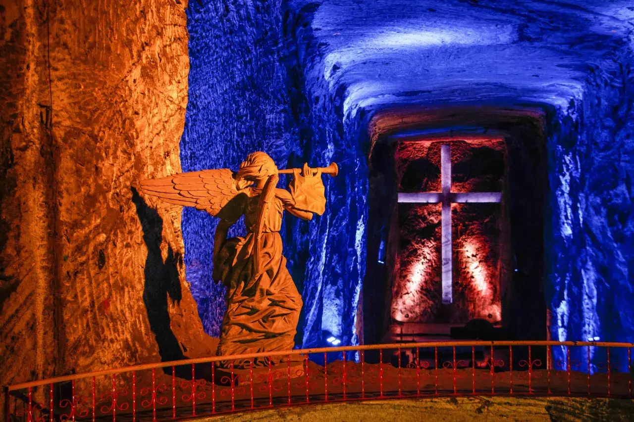 Statua di un angelo con tromba nella cattedrale del sale, illuminata da luci blu e arancioni, con una croce sullo sfondo.
