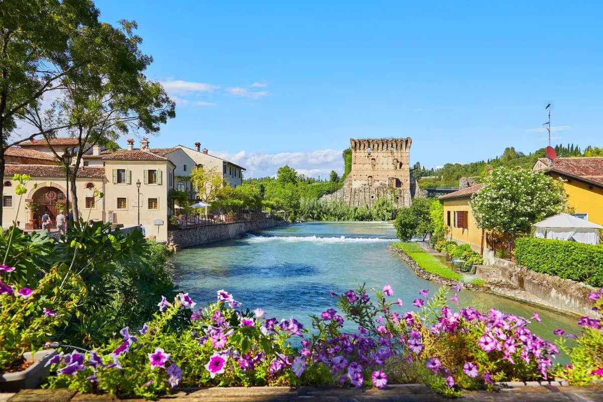 Un incantevole borgo da visitare in Veneto, con un fiume che scorre tra case antiche e una torre medievale. Fiori colorati incorniciano la scena.