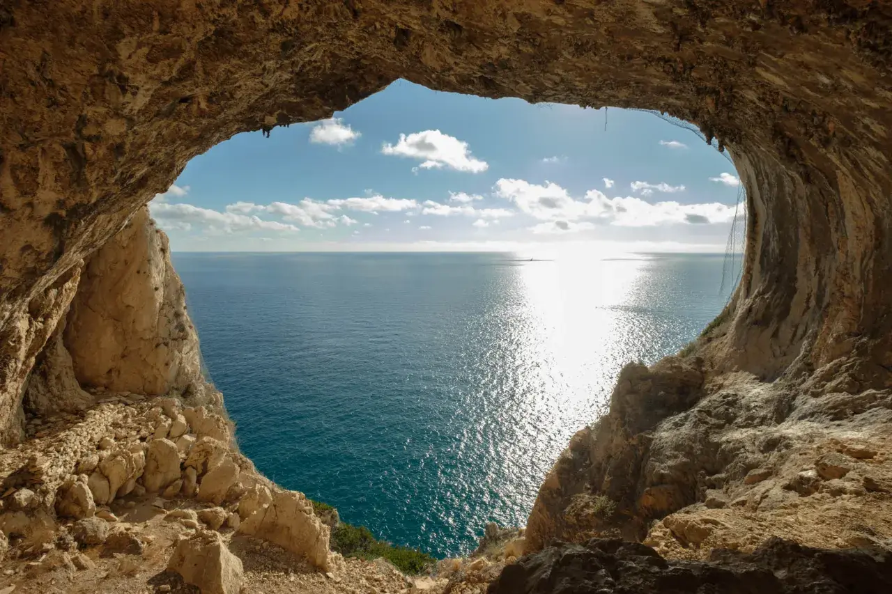 Vista dal mare da una grotta, con un sentiero del pellegrino che si intravede tra le rocce. Il sole brilla sull'acqua.