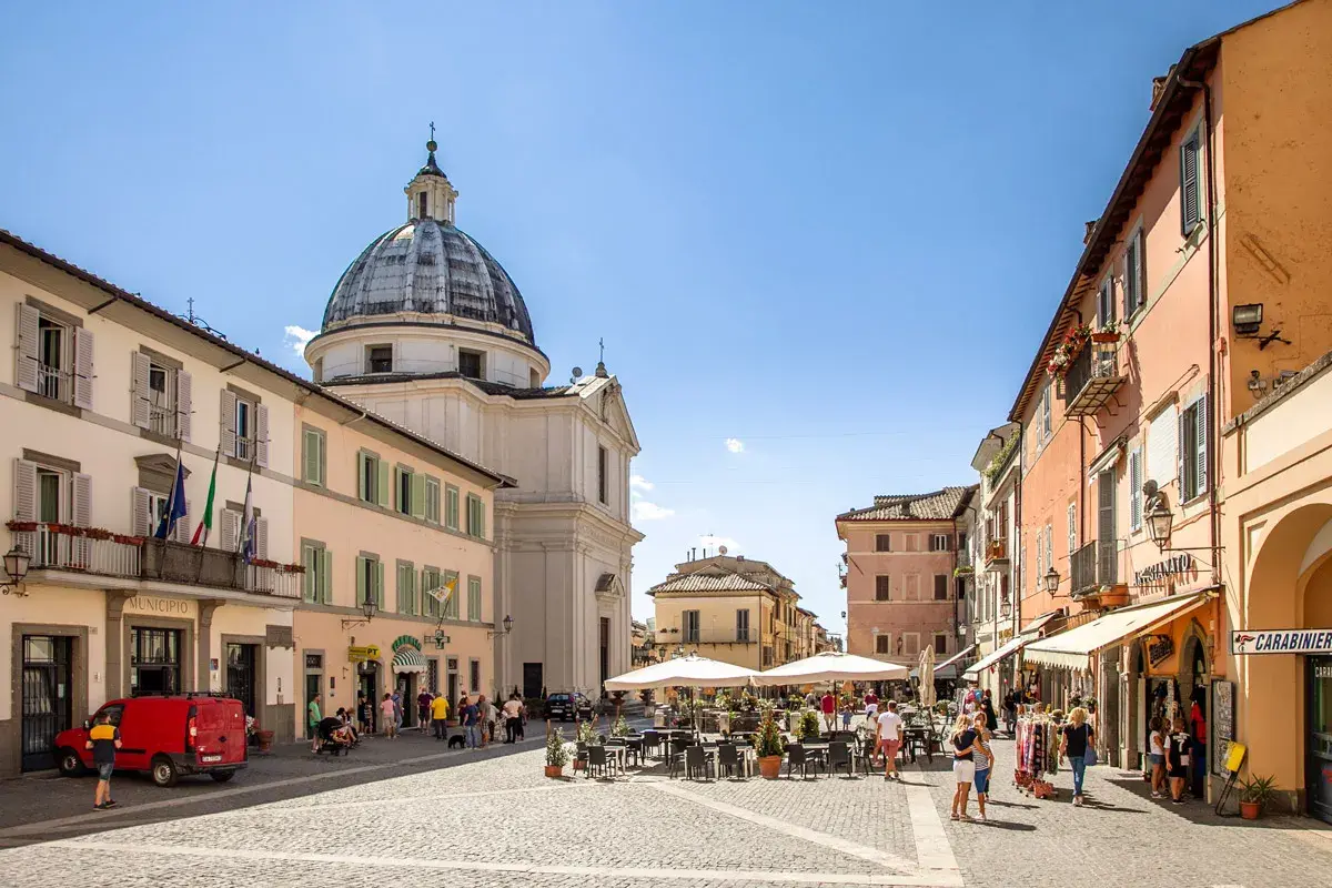 Piazza a Castel Gandolfo, dove si trova il Palazzo Apostolico. Edifici colorati, una chiesa con cupola e tavolini all'aperto animano la piazza.