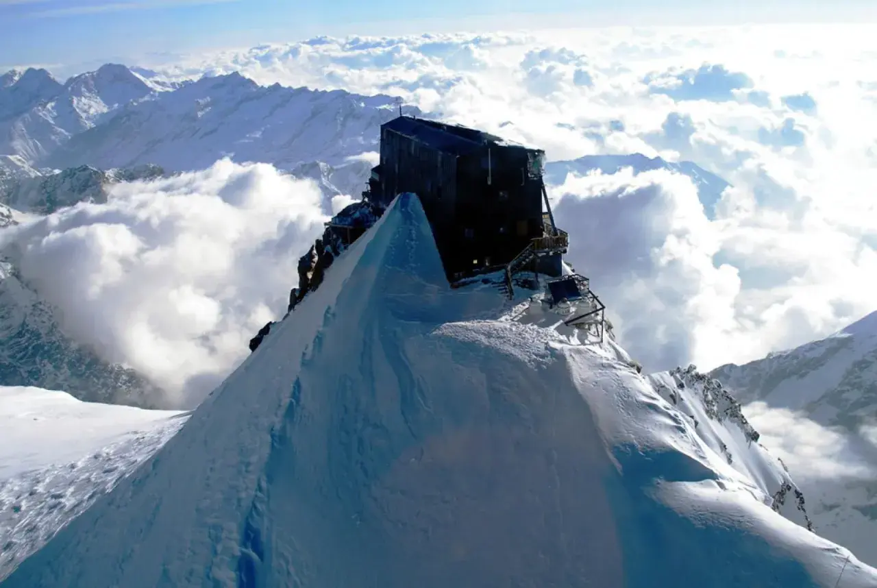 Il rifugio più alto d'Europa, un edificio scuro su una vetta innevata, domina un mare di nuvole e montagne.