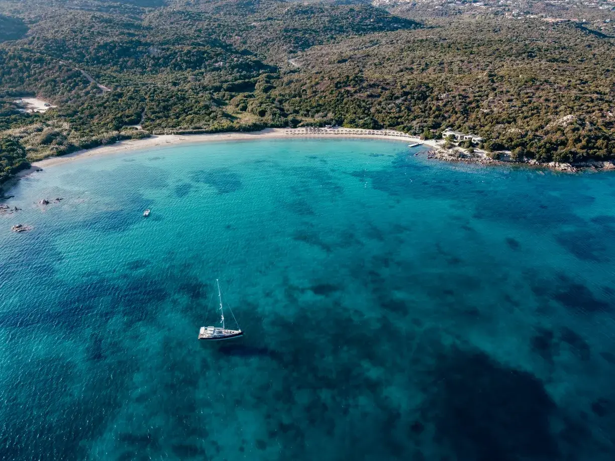 Vista aerea di una baia con acque cristalline, una barca a vela e una spiaggia dorata. Un assaggio delle migliori spiagge Costa Smeralda.