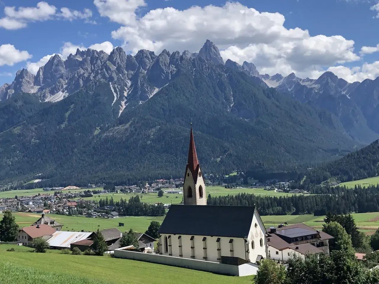Chiesa con campanile rosso in primo piano, sullo sfondo le maestose vette delle Dolomiti e i **paesi più belli** della **Val Pusteria**.