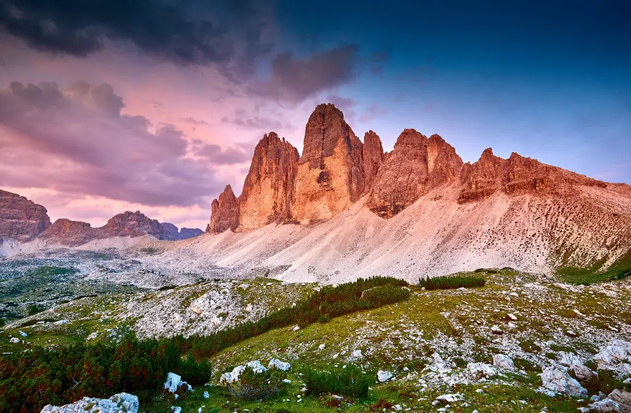Le maestose cime di Lavaredo illuminate dal tramonto, con un cielo viola e blu che fa da sfondo a un paesaggio alpino mozzafiato.
