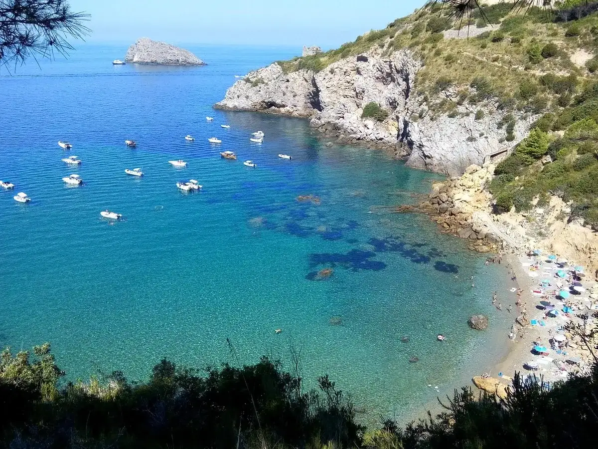 La cala del Gesso, Monte Argentario, offre un mare cristallino con barche all'ancora e una spiaggia affollata di bagnanti sotto ombrelloni colorati.
