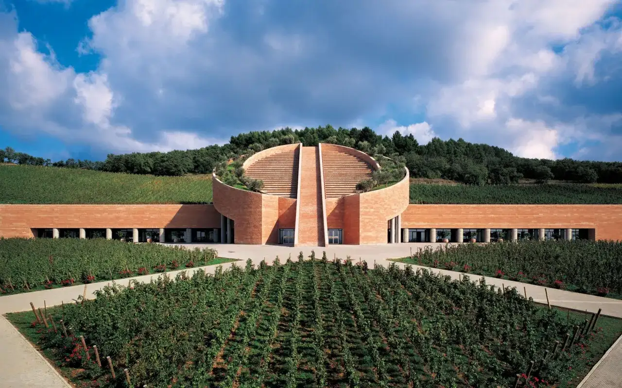 Cantine toscana famose: un edificio moderno in mattoni rossi, circondato da vigneti rigogliosi e alberi, con una scalinata semicircolare che sale verso il cielo.