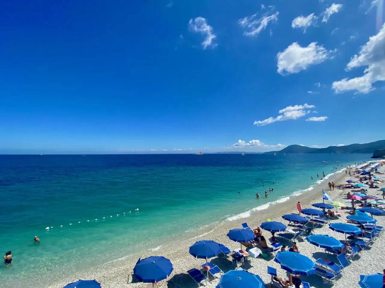 Spiaggia affollata nel golfo di Follonica, con ombrelloni blu e gente che si gode il mare cristallino sotto un cielo azzurro.