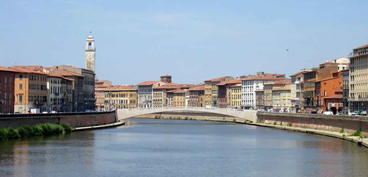Il fiume Arno attraversa il centro di Pisa, fiancheggiato da palazzi colorati e un ponte bianco. In lontananza, si staglia un'alta torre con un orologio.