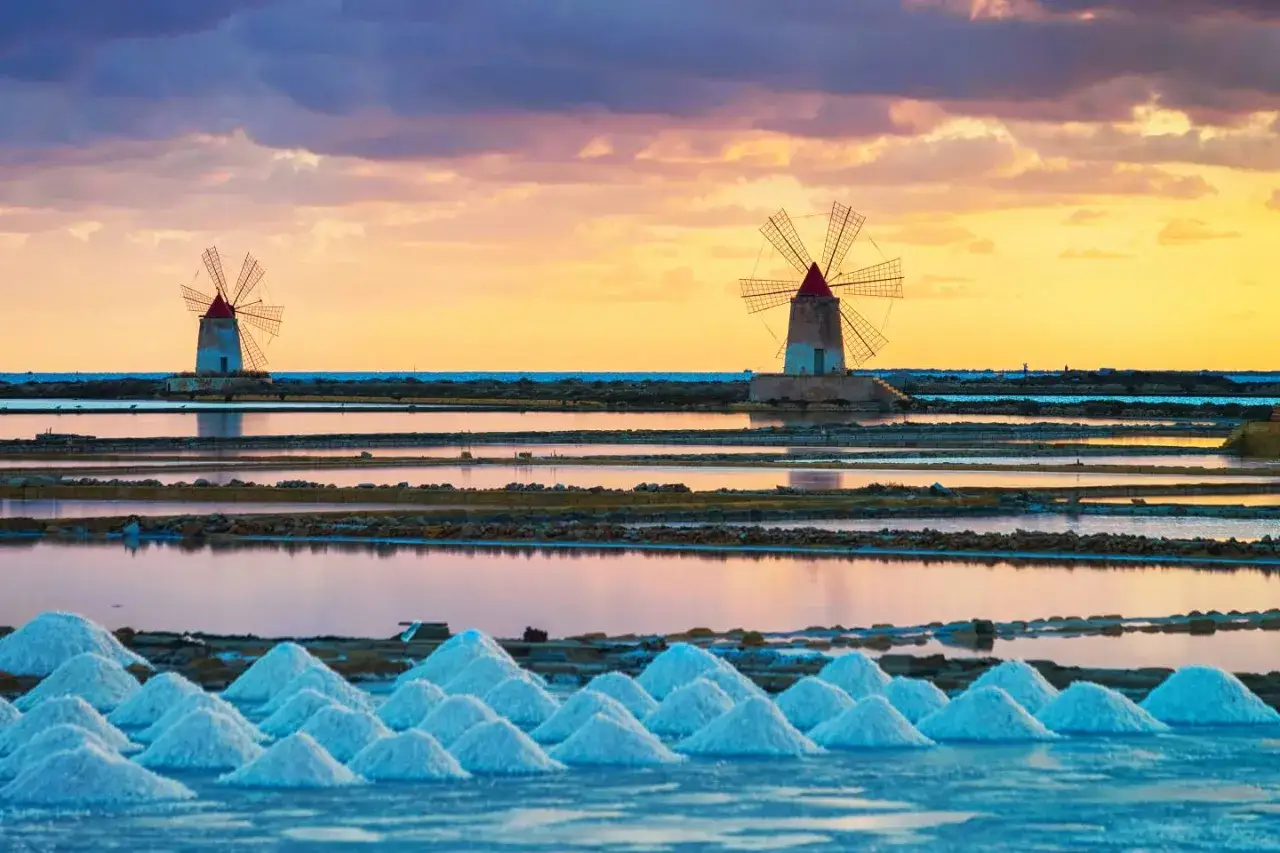 Saline di Trapani al tramonto, con mulini a vento e cumuli di sale. Un'idea di cosa vedere a Trapani in un giorno.