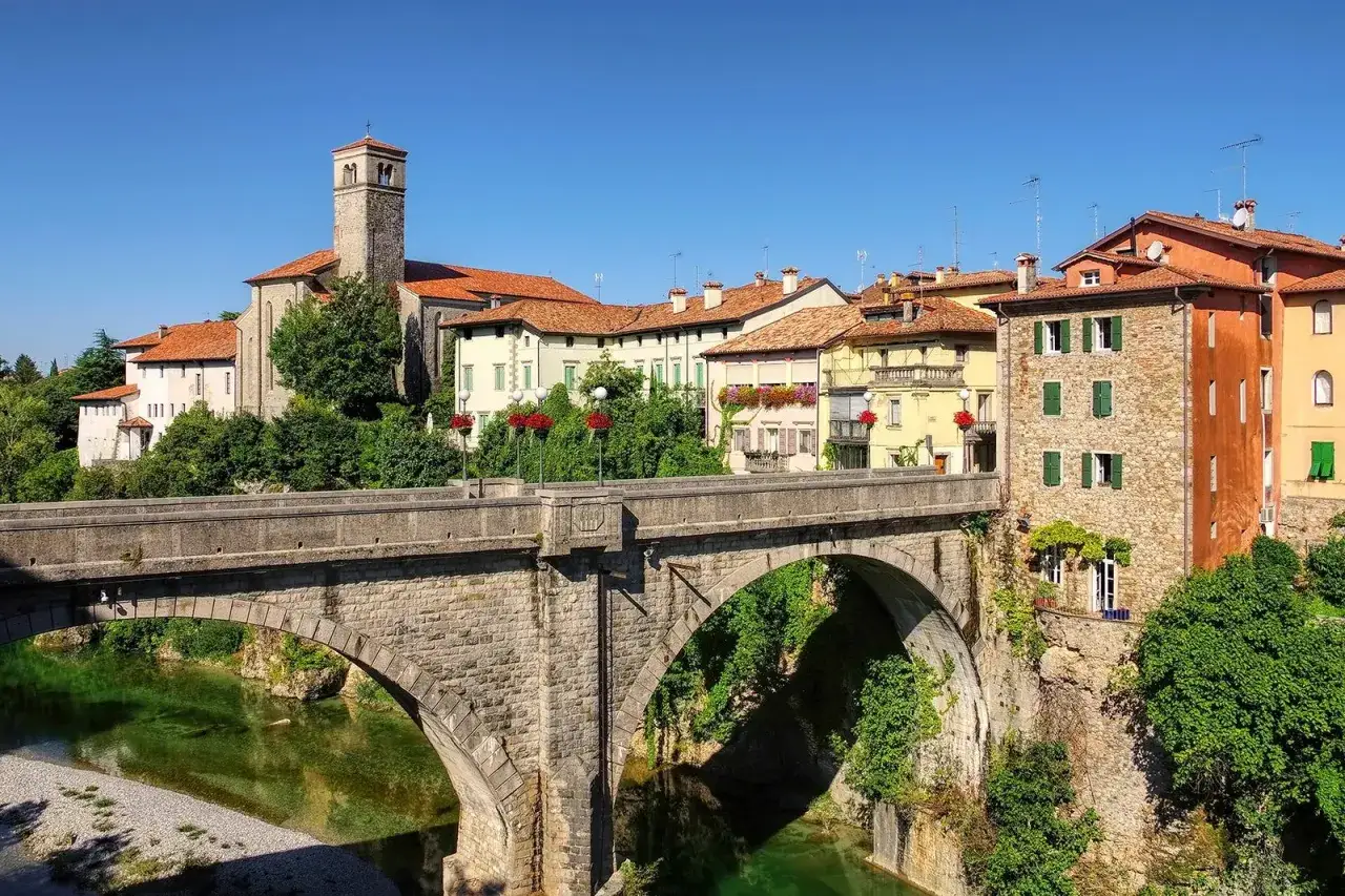 Ponte in pietra su fiume verde, con case colorate e campanile. Un gioiello tra i borghi Friuli da visitare.