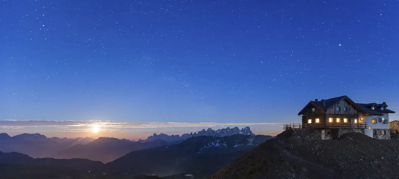 Rifugio illuminato su una montagna di notte, sotto un cielo stellato e con la luna che sorge all'orizzonte.