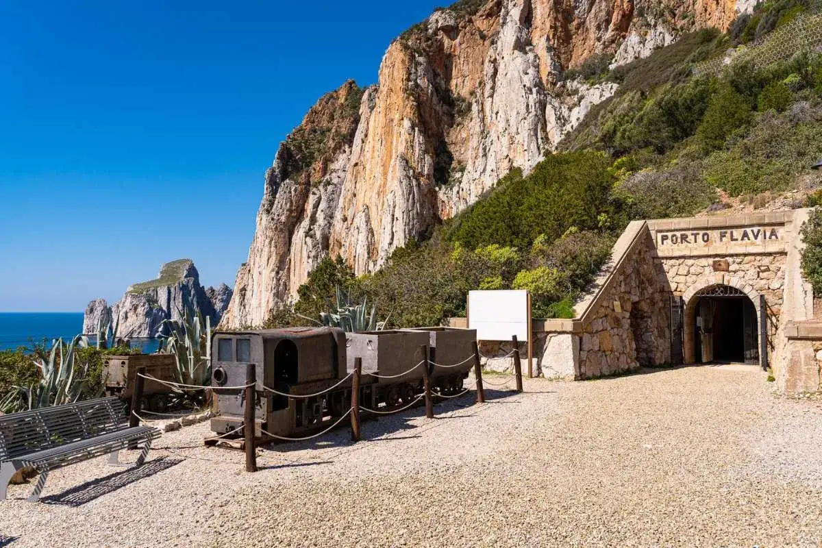 Antichi vagoni minerari e l'ingresso di Porto Flavia, testimoni della storia della zona mineraria sarda, si stagliano contro le imponenti scogliere.
