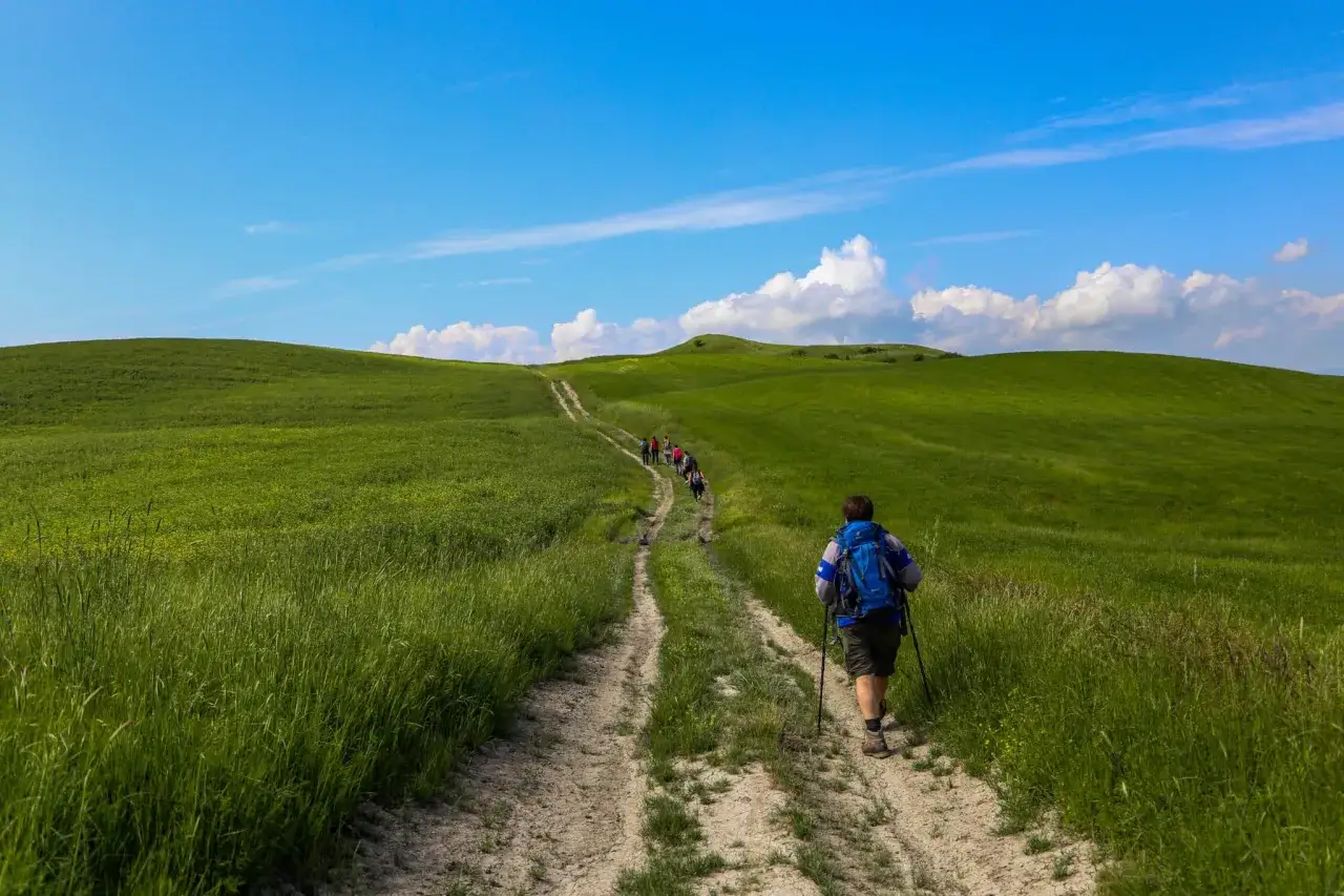 Un gruppo di escursionisti percorre un sentiero sterrato tra dolci colline verdi, sotto un cielo azzurro con nuvole bianche. Un'avventura lungo la via Lauretana Toscana.