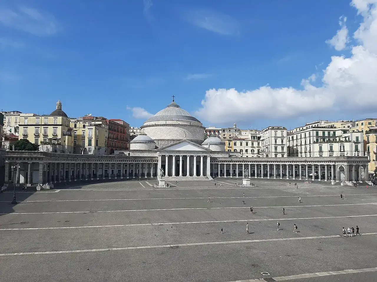 La maestosa piazza Plebiscito a Napoli, con il suo colonnato semicircolare e la Basilica di San Francesco di Paola, sotto un cielo azzurro con nuvole bianche.