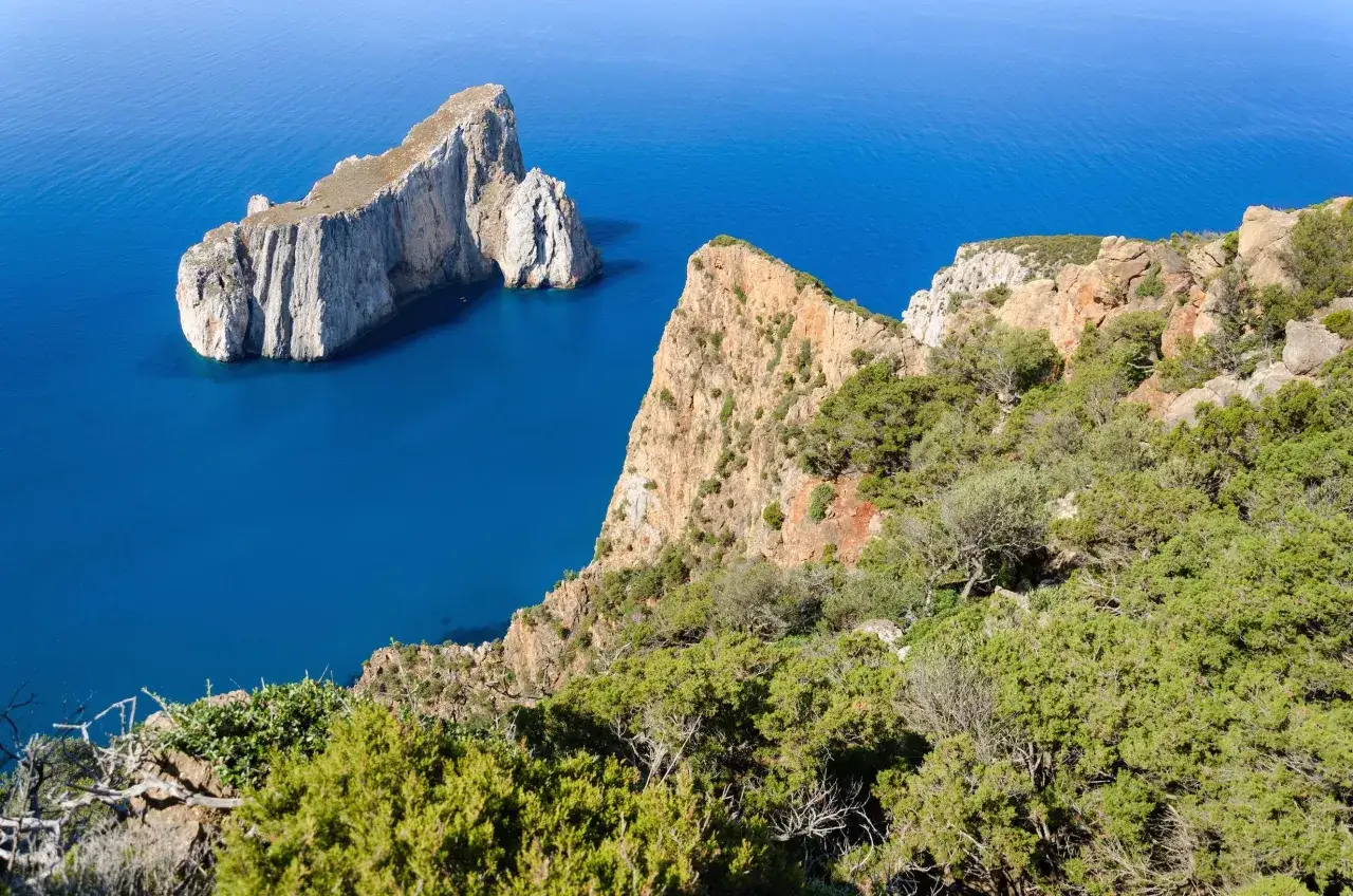 Scogliere rocciose e vegetazione mediterranea sulla costa ovest Sardegna, con il mare blu intenso che lambisce i faraglioni.