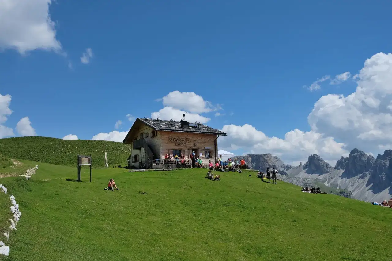 Un rifugio in Val Gardena, con persone che si rilassano sull'erba verde sotto un cielo azzurro e nuvoloso.
