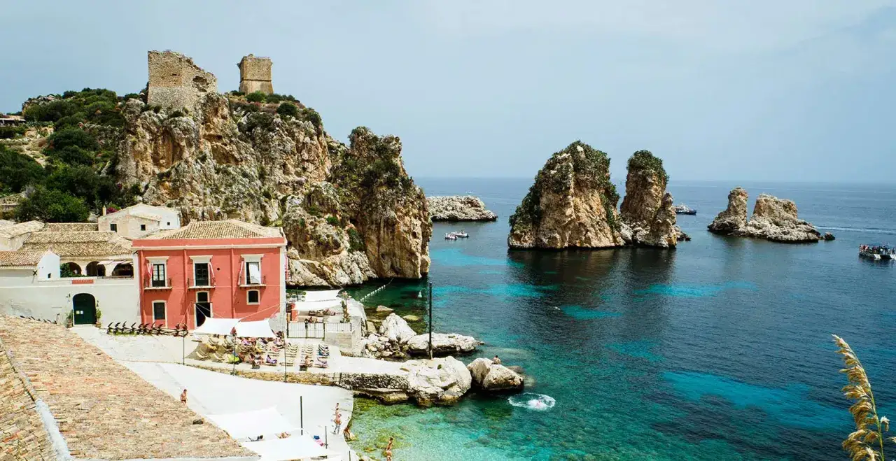 Scogliere rocciose e mare cristallino a Castellammare del Golfo, con case colorate e torri antiche.
