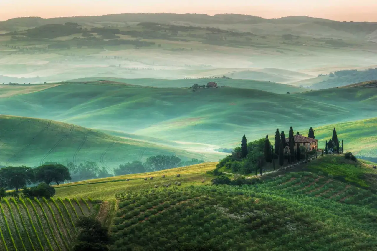 Dolci colline verdi della Toscana da vedere, con una casa colonica su un crinale e filari di cipressi.