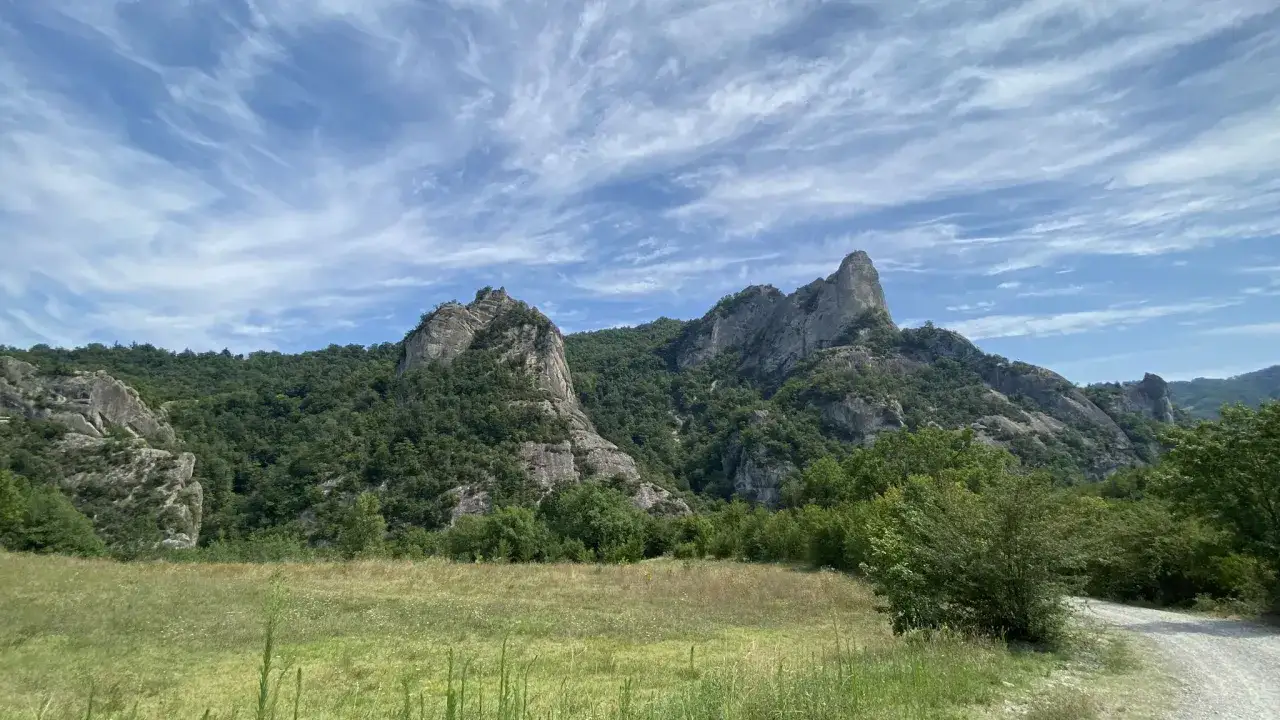 I Sassi di Roccamalatina, imponenti formazioni rocciose, emergono da una collina verdeggiante sotto un cielo azzurro solcato da nuvole bianche.