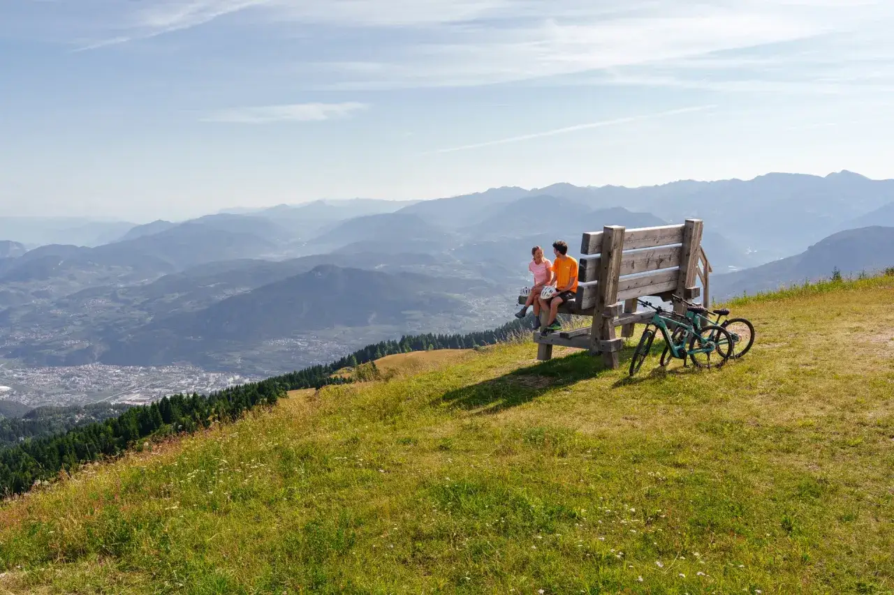 Coppia su panca con vista panoramica, ideale per passeggiate facili vicino a Trento. Due bici da corsa accanto, pronte per esplorare i sentieri.