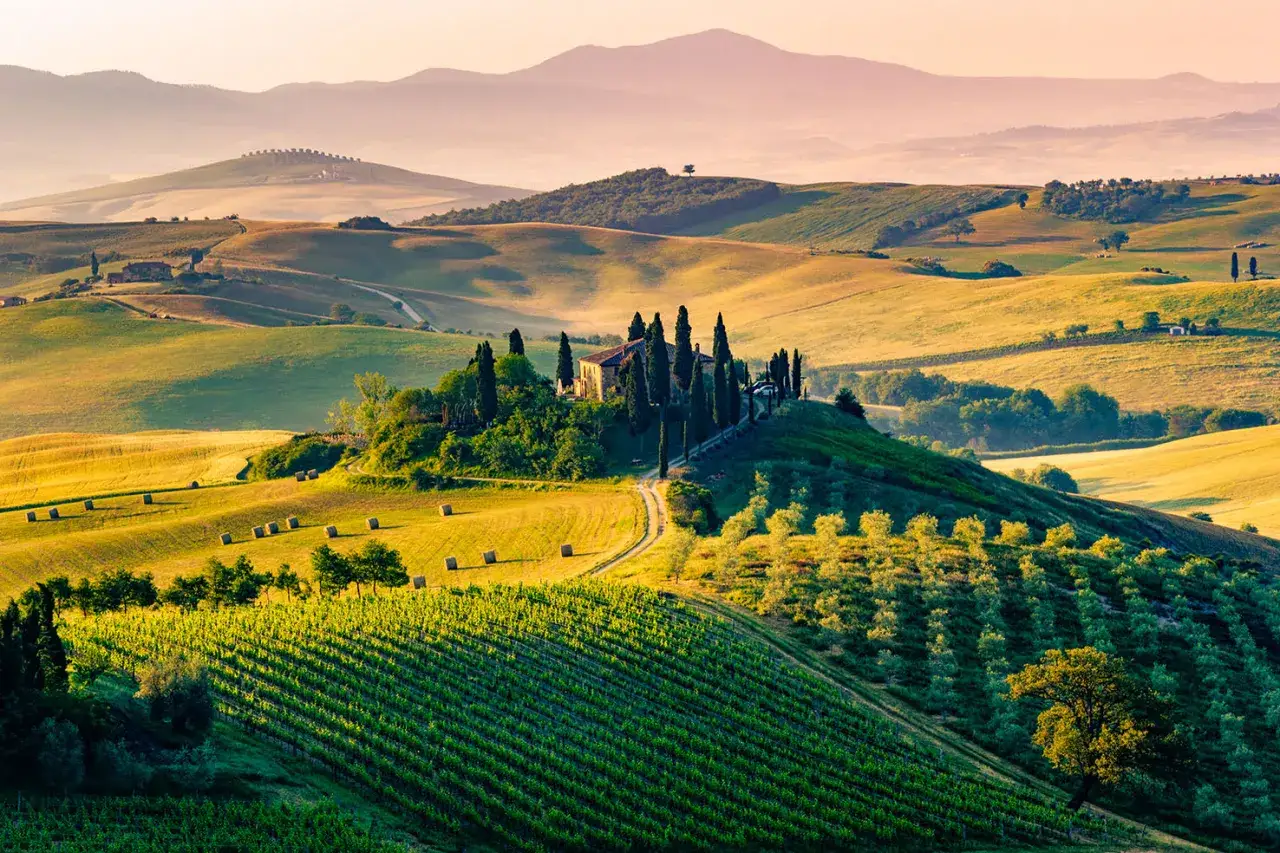 Colline ondulate con vigneti, balle di fieno e una casa in cima, un'immagine iconica dei borghi più belli Toscana.