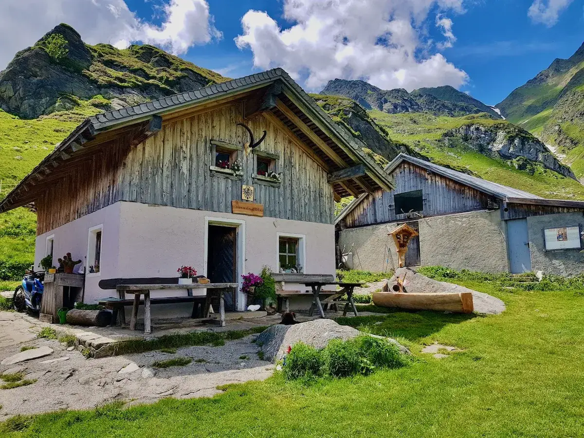 Rifugio alpino lungo il sentiero delle malghe, con tavoli in legno all'esterno e montagne verdi sullo sfondo.